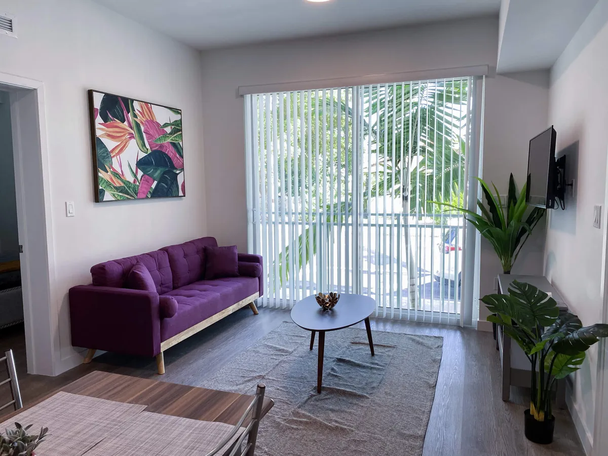 The image showcases a modern, sunlit living room with a plush purple sofa as the centerpiece, complemented by a stylish coffee table with a decorative centerpiece. Large vertical blinds let in abundant natural light, while lush tropical plants add a fresh, Miami-inspired vibe. The space feels open, cozy, and thoughtfully decorated, inviting guests to relax and feel at home.