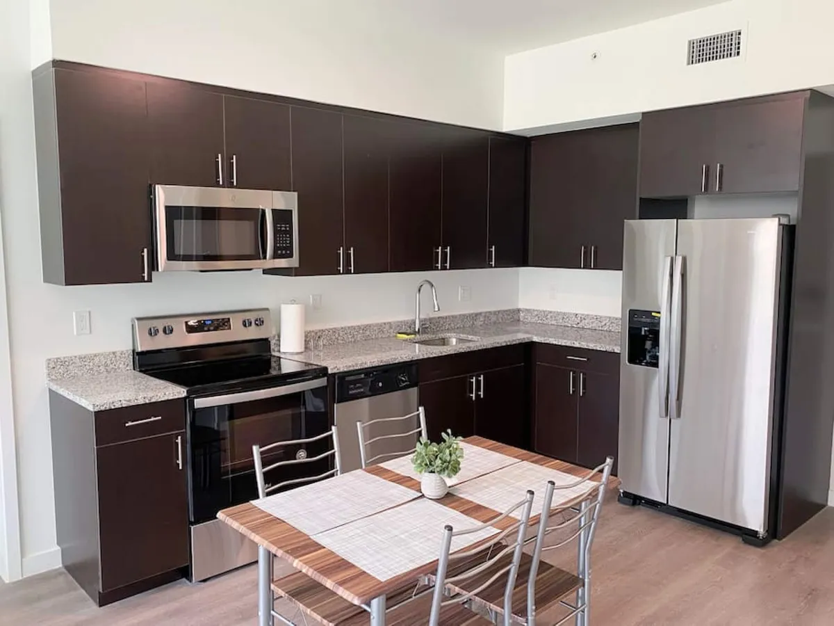 A bright, inviting kitchen space featuring stainless steel appliances, dark wood cabinetry, granite countertops, and a small dining table set for four with a plant centerpiece. The layout encourages both cooking and socializing, with ample space for meal prep and gathering.