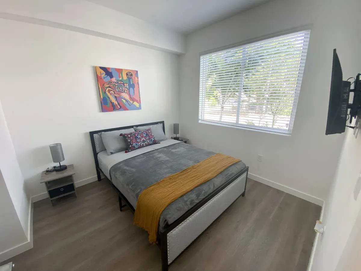 A tastefully decorated bedroom featuring a plush queen bed with gray bedding and a mustard throw. Above the bed hangs a colorful abstract painting, and the large window with white blinds allows natural daylight to brighten the space. A flat-screen TV is mounted on the wall, providing modern comfort.