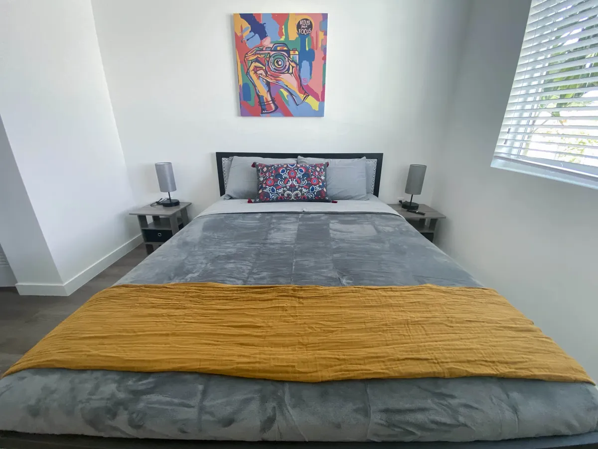 A cozy bedroom with a queen bed dressed in gray bedding and a mustard yellow throw. Colorful wall art and bedside tables with lamps flank the bed, while a window with blinds lets in ample natural light. The room is bright, relaxing, and inviting—ideal for rest and recovery.