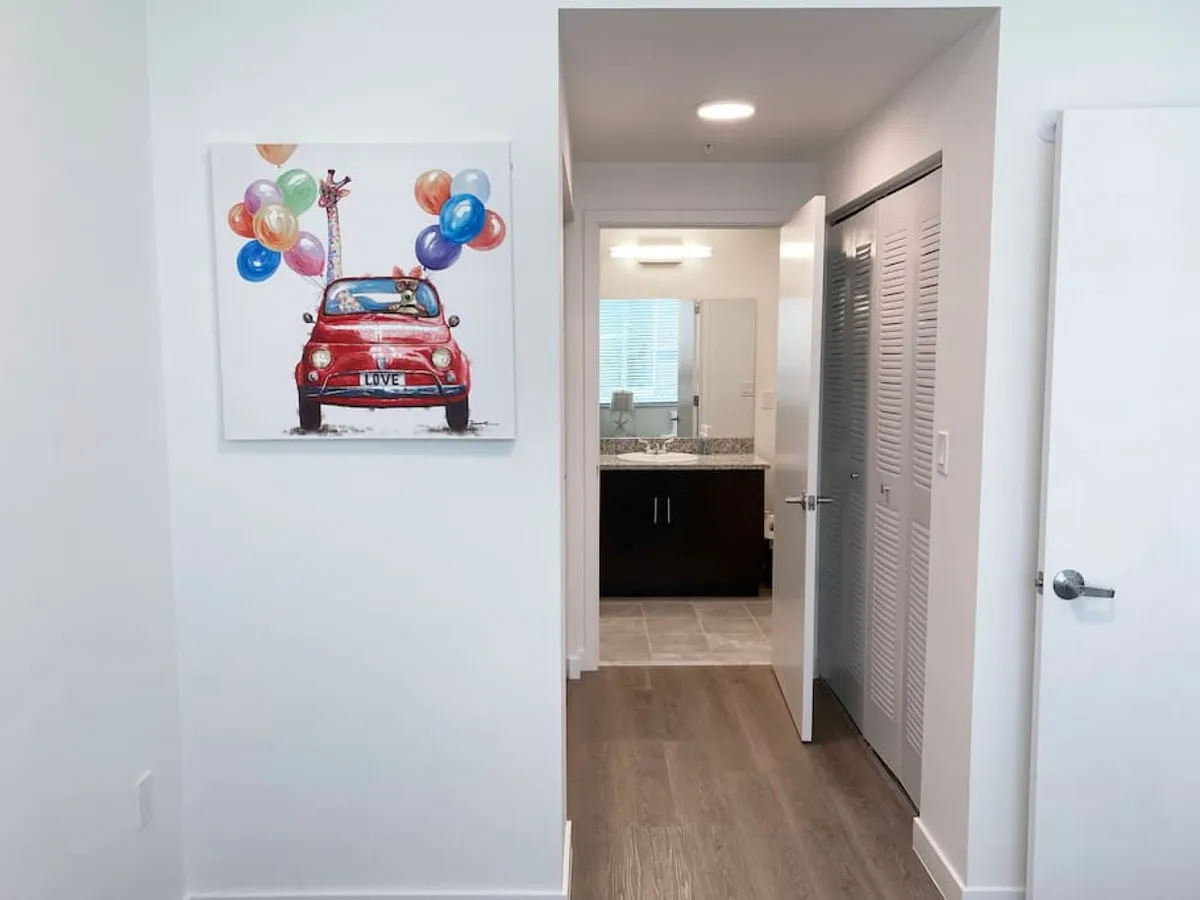 A bright interior hallway with wooden flooring, white walls, and doors leading to adjacent rooms. Colorful artwork featuring a red car and balloons adds a playful touch. The open doorway provides a view of a modern bathroom with a granite countertop, and the flooring continues seamlessly throughout, emphasizing the open and accessible layout.