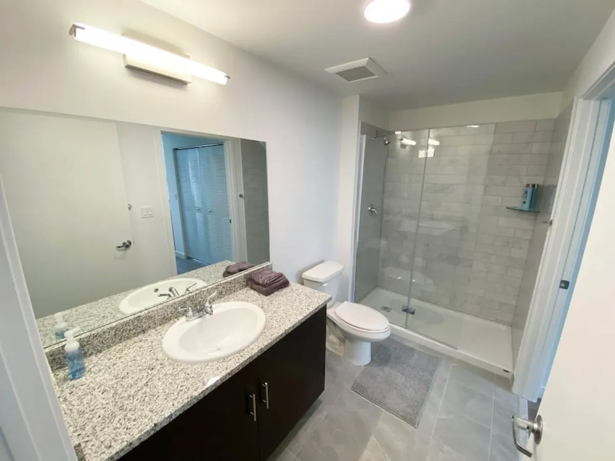 This image showcases a modern bathroom featuring a sleek granite countertop with a sink, a glass-enclosed shower, a toilet, and a large mirror above the sink. The environment appears clean, fresh, and calming, with design choices that promote both comfort and convenience. The space is accessible, with no stairs and thoughtful layout, making it suitable for a variety of guests, including those with mobility needs.