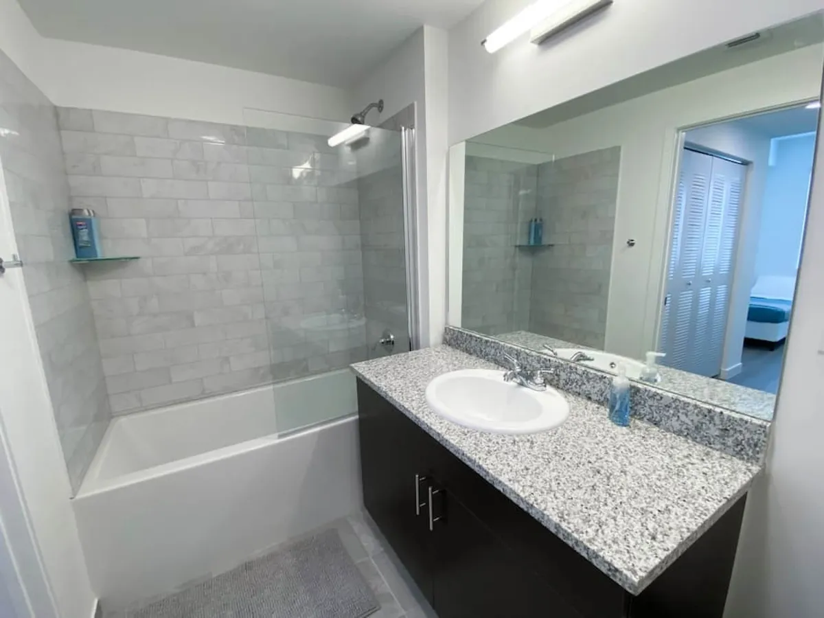 A modern bathroom featuring a granite countertop with a built-in sink, a large mirror, a bathtub with a tiled wall and glass partition, and a view toward the adjacent bedroom. The fresh, practical atmosphere is ideal for relaxation and refreshment.